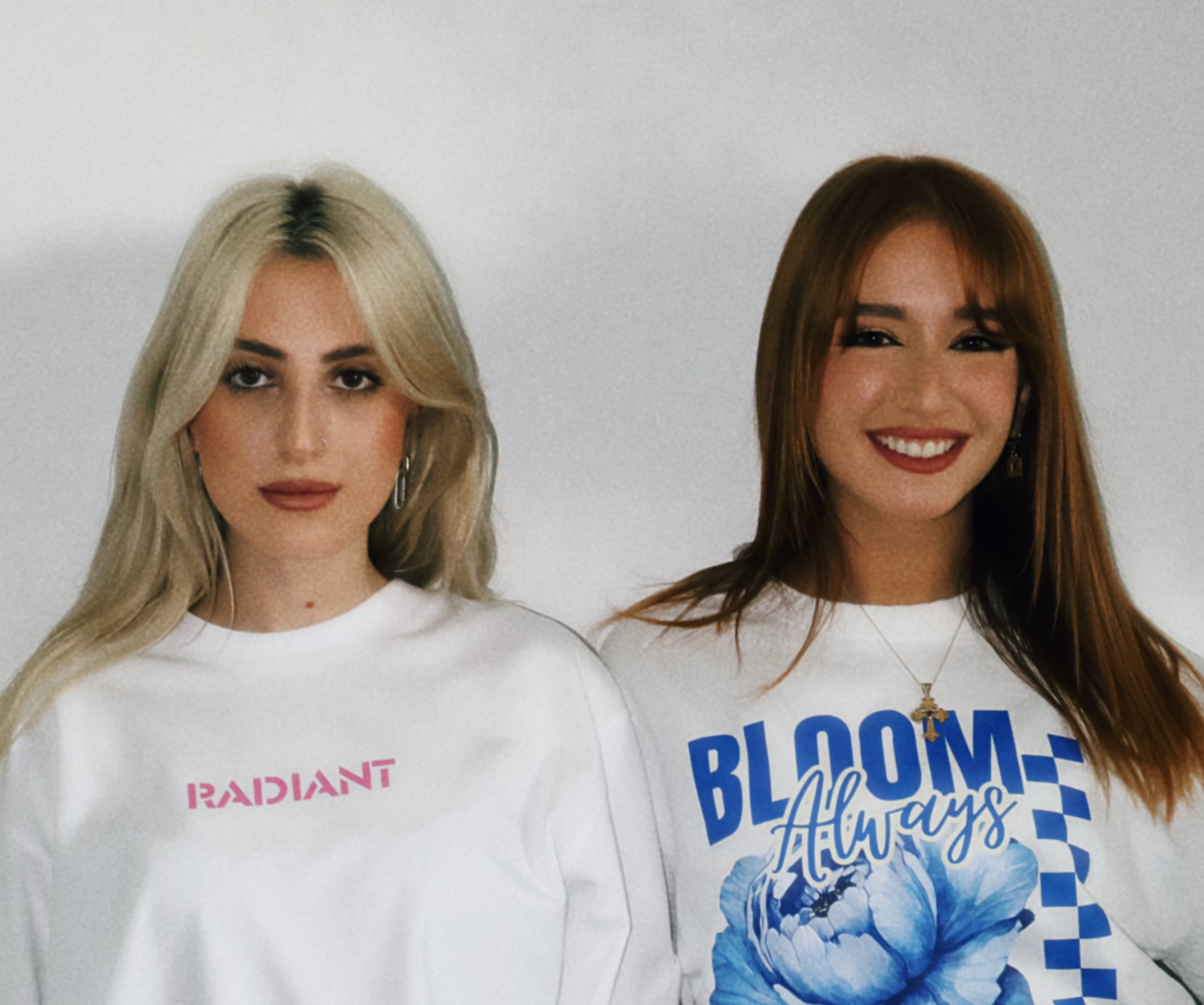 Two women wearing white t-shirts with text and a blue flower design Radiant Clothes Cyprus.